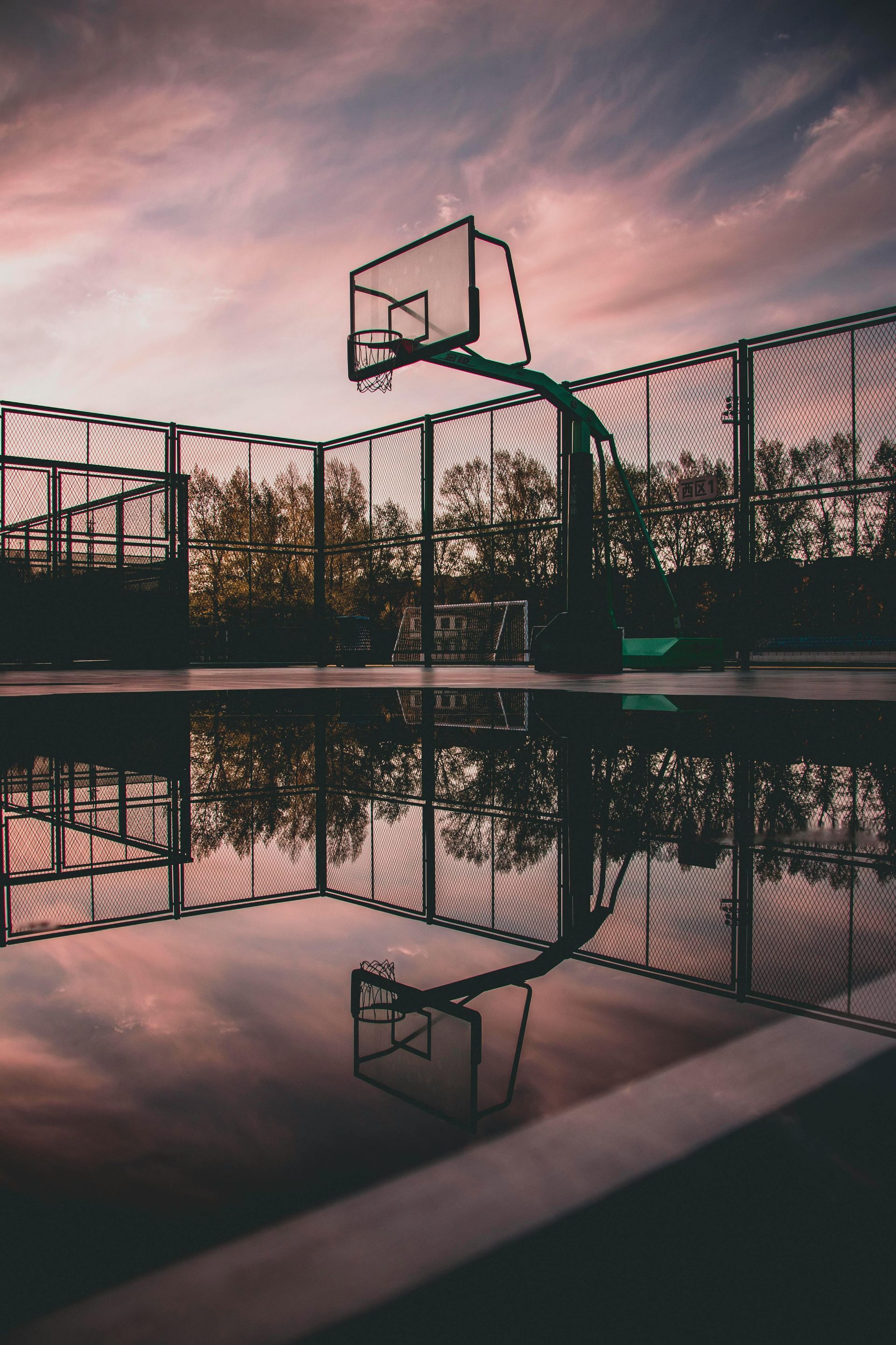 Empty outdoor basketball court at sunset, reflected in a puddle with pink sky above