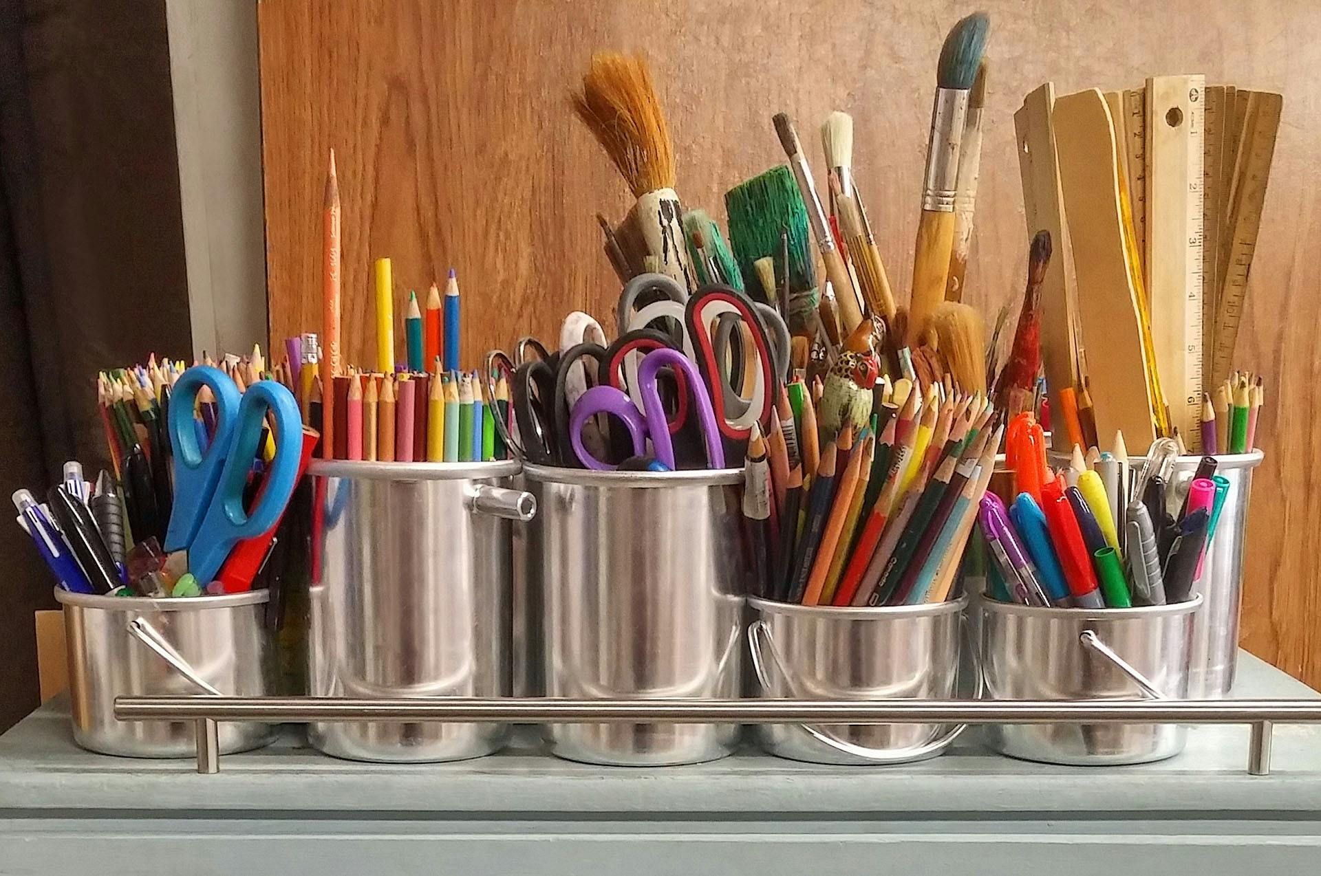 Assorted paintbrushes, pencils, and scissors in metal cups on a wooden shelf